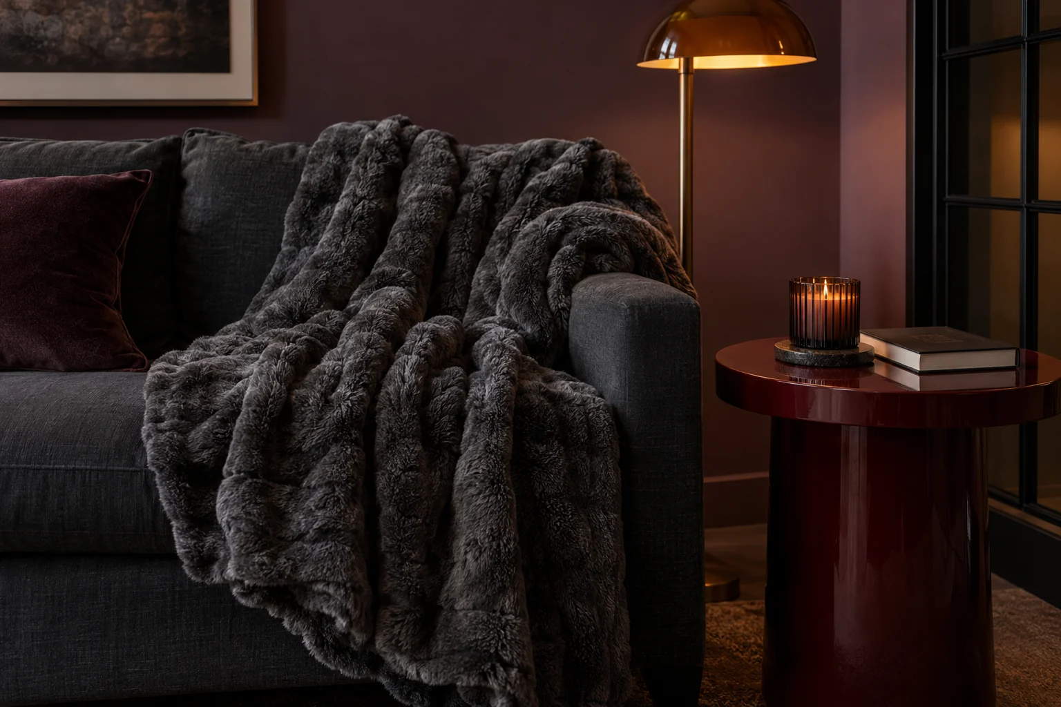 A charcoal sofa with a ruched dark-gray throw beside a candle and a burgundy side table in warm evening light